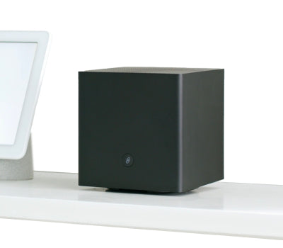 A black, cube-shaped electronic device on a white shelf, next to a screen.