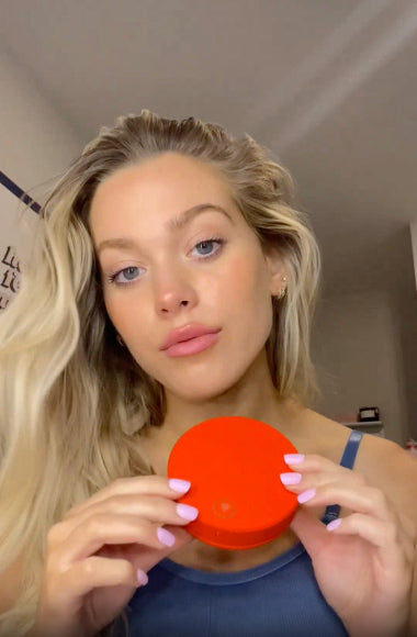 Person holding a red circular container, with long blonde hair, in a casual indoor setting.