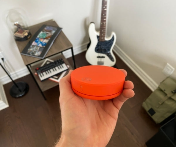 Hand holding an orange circular device; room with guitar and keyboard in background.